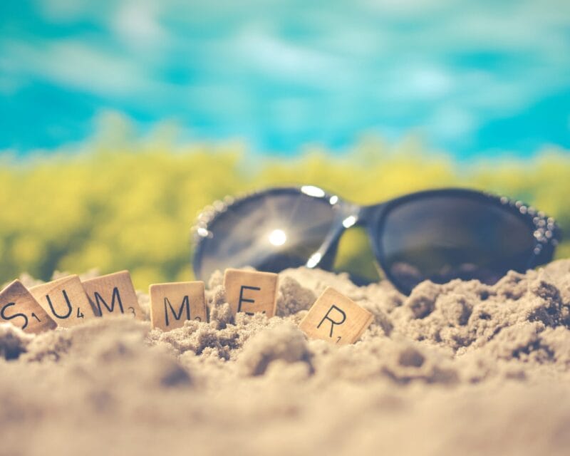 Scrabble letters spelling "Summer" on a beach with sunglasses in background